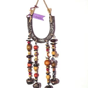 May include: A rustic horseshoe wall hanging with beaded strands and small bells. The horseshoe is dark brown with a jute rope hanger. Strands of wooden beads in various colors and shapes dangle below, with small metal bells at the bottom. A purple tag reads "Handmade".