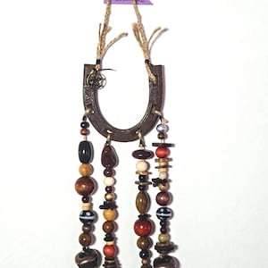 Puede incluir: Un colgante de pared rústico en forma de herradura con hilos de cuentas de madera de varias formas y colores, incluyendo marrón, negro y rojo. La herradura es de metal y tiene un pequeño dije de estrella. La palabra "handmade" está en una etiqueta morada.