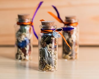 Puede incluir: Tres pequeñas botellas de vidrio llenas de hierbas y especias secas. Cada botella tiene un tapón de corcho, atado con cintas naranjas y moradas. Un dije plateado cuelga de la botella central. Las botellas están dispuestas sobre una superficie de madera clara.