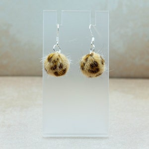 May include: A pair of dangle earrings with a leopard print pom-pom charm. The earrings are silver and the pom-poms are brown and white.