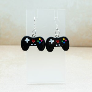 May include: A pair of black video game controller earrings with colorful buttons. The earrings are hanging from silver hooks.
