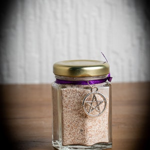 Może przedstawiać: Szklany słoik ze złotą pokrywką i fioletową wstążką przewiązaną wokół. Słoik zawiera biały proszek i ma srebrny wisiorek w kształcie pentagramu wiszący na wstążce.