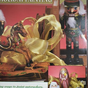 May include: A still life featuring a wooden rocking horse, a nutcracker, and nesting dolls. The scene is decorated with gold ribbon and books. The text "HOLIDAY PAINTING" is visible. The image has a festive, holiday theme.