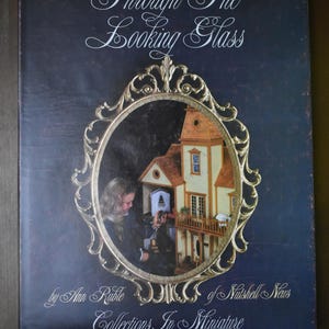 May include: A dark blue book titled Through The Looking Glass by Ann Ruble of Nutshell News Collections In Miniature. The cover features an ornate gold-colored oval frame surrounding a photograph of a person looking at a miniature dollhouse.