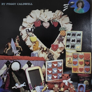 May include: A book cover titled "BASIC BRUSH STROKES on Wooden Cut Outs" by Peggy Caldwell. The cover features a heart-shaped wreath with various wooden cutouts, including apples, hearts, and a gingerbread man. Other folk art items are also displayed.