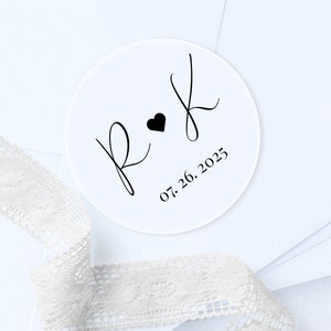 May include: A white round sticker with black calligraphy initials "R & K" with a small heart between the letters and the date "07.26.2025" below.