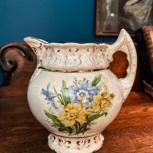 May include: A cream-colored ceramic pitcher featuring a floral decal of blue and yellow daffodils with green leaves. The pitcher has a scalloped rim, a textured base, and a gold-painted trim along the center and the handle edges.