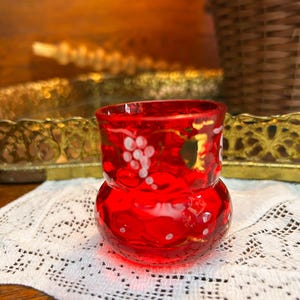 May include: A small, vibrant red glass votive candle holder. The candle holder features a bulbous base and a slightly flared top. White grape cluster designs and gold accents adorn the exterior. The candle holder sits on a white lace doily.