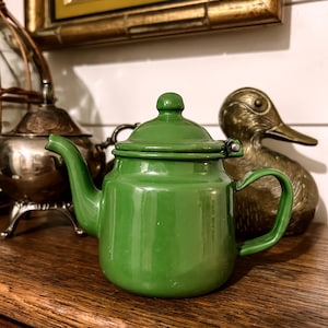 May include: A vintage, bright green enamel teapot with a matching lid and handle. The teapot is set on a wooden surface, with a brass duck figurine and a silver tea set in the background. The teapot has a classic, rounded shape.