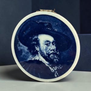 May include: A round, blue and white ceramic plate featuring a portrait of a man in a hat. The portrait is in a detailed, illustrative style. The plate has a white border and a small loop at the top.
