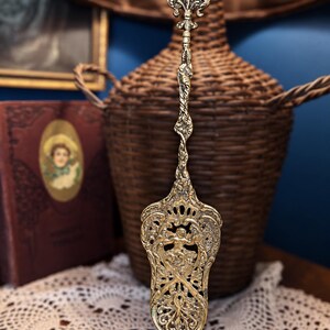 May include: A gold-toned metal cake server featuring an ornate, pierced handle with a fleur-de-lis finial and a wide, flat blade decorated with intricate scrollwork and a central cherub motif, showcasing a detailed, embossed metallic finish.