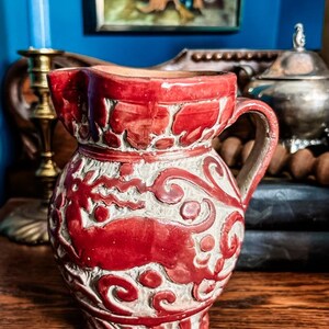 May include: A ceramic pitcher featuring a glossy red glaze over a textured off-white base. The surface is decorated with raised relief patterns depicting a stylized animal figure surrounded by swirling scrollwork and geometric border designs.
