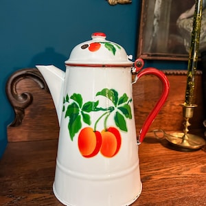 May include: White enamelware coffee pot with a red handle and lid trim. The pot is decorated with green leaves and orange fruit. A vintage kitchenware item.