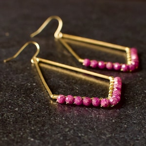 May include: A pair of gold earrings with a geometric shape and pink gemstone accents.