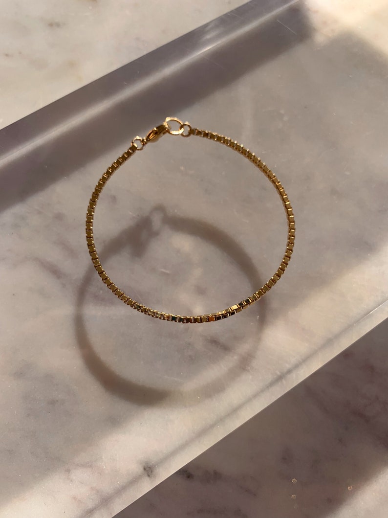 May include: A gold chain bracelet with a delicate box chain design. The bracelet is made of a thin, delicate chain and has a simple clasp.