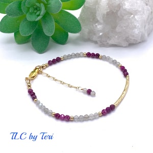 May include: A gold beaded bracelet with a gold bar charm. The beads are a mix of white and purple.