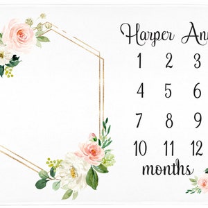 May include: A white baby blanket with a gold geometric frame and pink floral accents. The text "Harper Ann" and "1 2 3 4 5 6 7 8 9 10 11 12 months" is printed on the blanket.