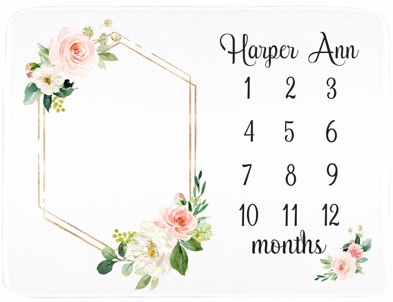 May include: A white baby blanket with a gold geometric frame and pink floral accents. The text "Harper Ann" and "1 2 3 4 5 6 7 8 9 10 11 12 months" is printed on the blanket.