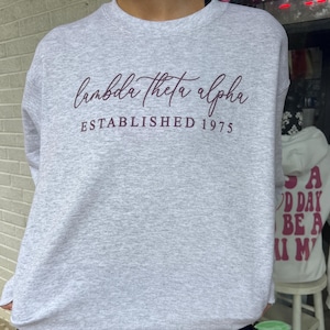 May include: A gray sweatshirt with the text "lambda theta alpha established 1975" in maroon lettering.