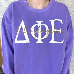 May include: Purple sweatshirt with white Greek letters that spell out "Delta Phi Epsilon" in a gold script font.
