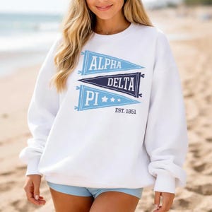 May include: White sweatshirt with blue text and pennant designs. The sweatshirt features the words "ALPHA DELTA PI" and "EST. 1851". The design is in a pennant style with blue and navy blue colors. The sweatshirt is a crew neck style.