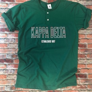 May include: A dark green t-shirt with a white button-up collar and a white graphic that says "Kappa Delta Established 1897".