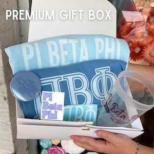 May include: A white gift box filled with Pi Beta Phi themed items, including a blue and white t-shirt, a blue button with the letters "Pi Beta Phi", a sticker with the letters "Pi Beta Phi", and a clear tumbler with the letters "Pi Beta Phi" and the text "Est. 1867".