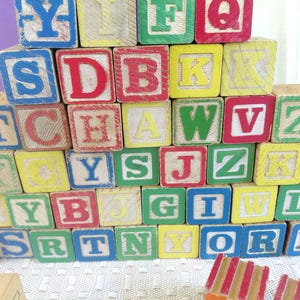 May include: A large pyramid stack of square wooden alphabet blocks featuring various letters in red, blue, green, and yellow paint. The blocks show signs of wear and are arranged in multiple rows, displaying a mix of uppercase letters on each face.