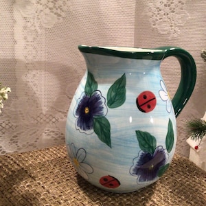 May include: A blue and white ceramic pitcher with a green rim and handle. The pitcher is decorated with painted flowers, leaves, and ladybugs.