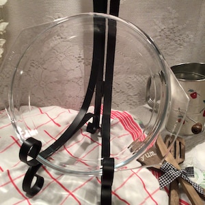 Puede incluir: Un molde para tartas de vidrio transparente con un soporte de metal negro. El soporte tiene un diseño decorativo de desplazamiento. El molde para tartas está sobre un mantel a cuadros rojos y blancos.