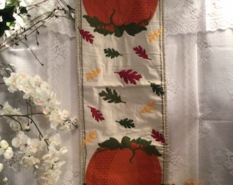 May include: A rectangular autumn-themed table runner featuring two large orange pumpkins and colourful leaves. The pumpkins are surrounded by green leaves and the runner has a cream background. The runner is hanging on a clear hanger.
