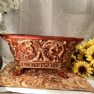 May include: A decorative planter with ornate gold and red detailing. The planter has four feet and is made of ceramic.