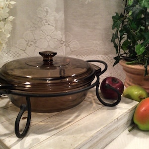 Puede incluir: Una fuente de vidrio marrón con tapa, colocada en un soporte de metal negro. El plato se encuentra sobre una superficie de madera blanca. Frutas decorativas y vegetación en el fondo.