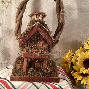 May include: A rustic wooden birdhouse with a moss-covered roof and a twig wreath around the top. The birdhouse is sitting on a red and white checked tablecloth.