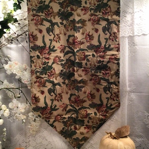 May include: A beige table runner with a floral pattern in shades of red, green, and brown. The runner has a tassel at the end and is hanging from a hanger.
