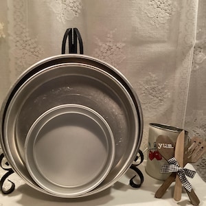 May include: Three round, silver-colored baking pans of varying sizes are stacked on a black metal stand. A mug with cherry graphics and the word "yum" is next to wooden utensils tied with a black and white checkered ribbon.
