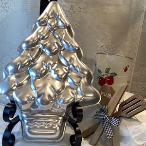 Puede incluir: Molde para pastel de árbol de Navidad de metal plateado con soporte. El molde tiene forma de árbol de Navidad con una superficie texturizada. El soporte es de metal negro con un diseño decorativo de desplazamiento.