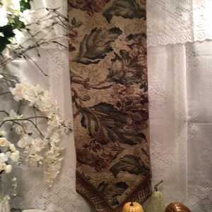Op de afbeelding: Een bruine, groene en beige tapijt tafelkleed met een bloemenpatroon. De tafelkleed heeft franjes aan de uiteinden en hangt aan een metalen staaf. De tafelkleed wordt getoond op een witte kant tafelkleed met drie decoratieve pompoenen.