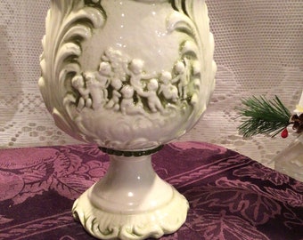 May include: White ceramic planter with a green glaze. The planter features a raised design of cherubs playing music. The planter has a pedestal base.