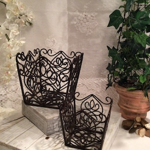 May include: Two decorative metal plant pots with intricate scrollwork designs. The pots are brown and have a square shape. They are sitting on a white wooden surface.