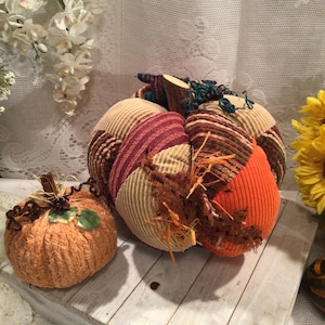 May include: Two fabric pumpkins, one large and one small. The large pumpkin is made of patchwork fabric in shades of orange, brown, and cream. The small pumpkin is a solid orange color. Both pumpkins have a fuzzy texture.