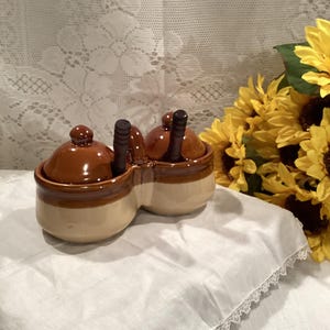 May include: A brown and beige ceramic double condiment dish with two wooden spoons. The dish is shaped like a peanut and has two separate compartments for holding condiments.