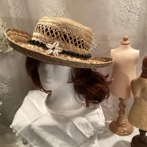 May include: A wide-brimmed straw hat with a woven design and a black beaded trim. A white flower made of seashells is attached to the hat.
