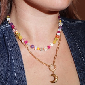 May include: A layered necklace with a gold chain and a beaded necklace. The gold chain has a crescent moon pendant. The beaded necklace features colorful beads in various shapes and sizes. The person is wearing a denim jacket and flower-shaped earrings.