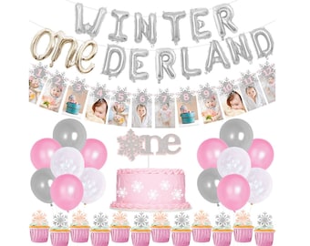 May include: Winter Wonderland first birthday party decorations with silver and pink balloons, a cake with snowflakes, cupcakes with snowflake toppers, and a banner with photos and the numbers 1-12.