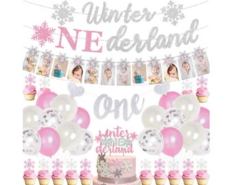 May include: Winter Wonderland themed first birthday party decorations with a silver glitter banner that says "Winter Wonderland" and a pink glitter banner that says "One". There are also silver and pink snowflake decorations, a cake with a silver and pink snowflake design, and a banner with photos of a baby girl.