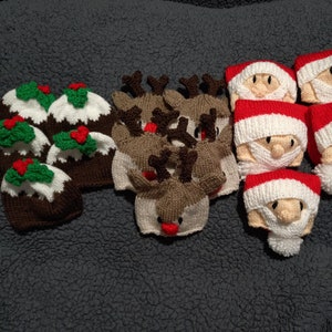 Könnte beinhalten: Eine Sammlung handgestrickter Weihnachtsmützen. Die Mützen haben die Form von Weihnachtsmann, Rentier und Weihnachtspudding. Die Mützen sind alle aus verschiedenen Garnfarben gefertigt, darunter Rot, Weiß, Grün, Braun und Schwarz.