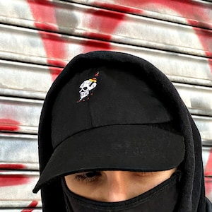 May include: A person wearing a black hooded sweatshirt and a black baseball cap with a white skull and crossbones graphic. The person is wearing a black face mask.