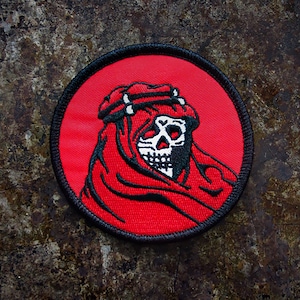 Pode incluir: Um remendo bordado vermelho com contorno preto, representando uma caveira usando um turbante vermelho e preto. A caveira tem olhos e dentes brancos.