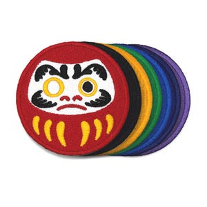 May include: A stack of round embroidered patches featuring a Daruma doll design. The patches are in various colors, including red, black, yellow, green, blue, and purple. The Daruma doll has white facial features and yellow eyes.